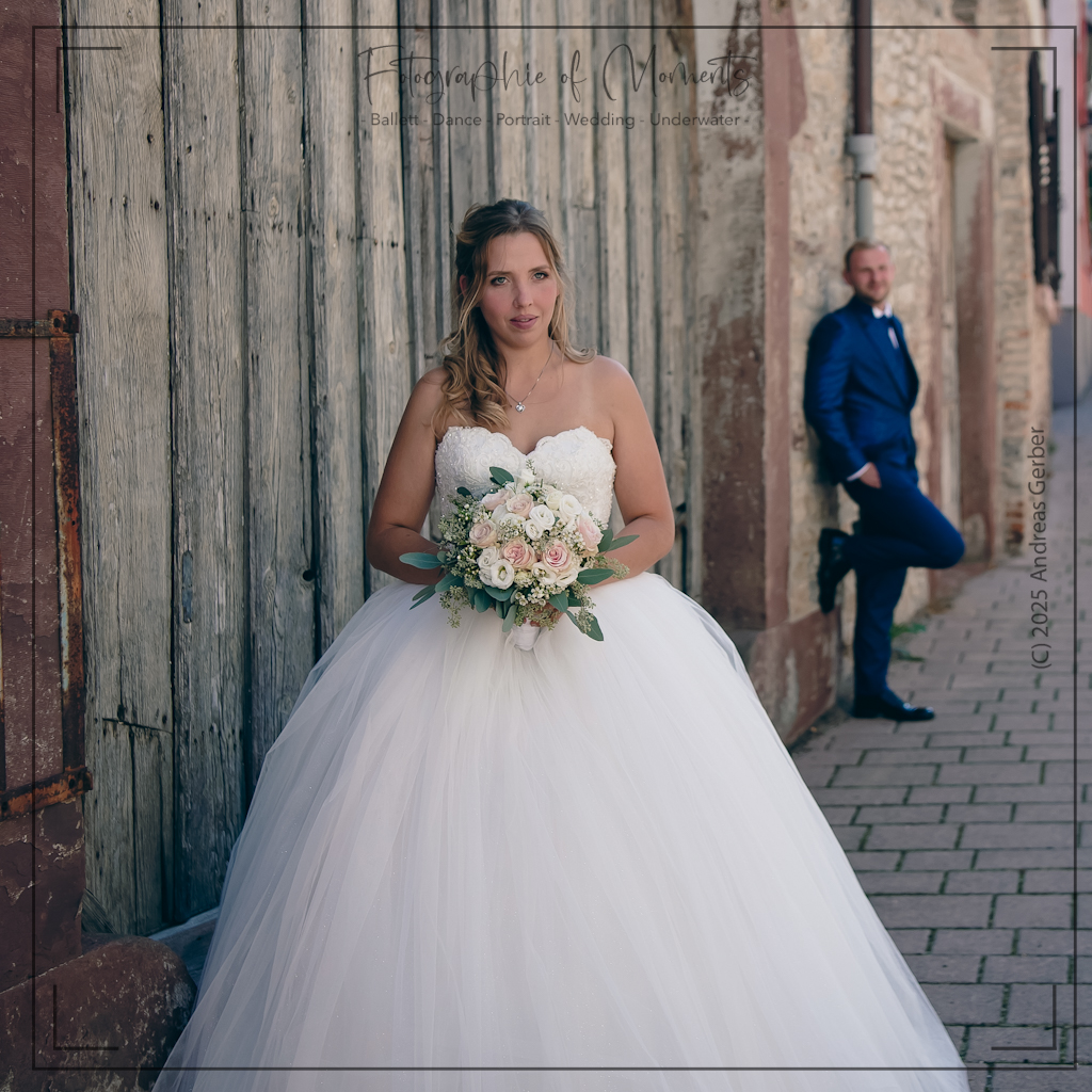 Braut mit Brautstrauß, Bräutigam lehnt an Steinwand.